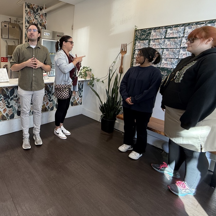 Students listen to a speaker inside a small café as an interpreter, Nichol signs beside him.