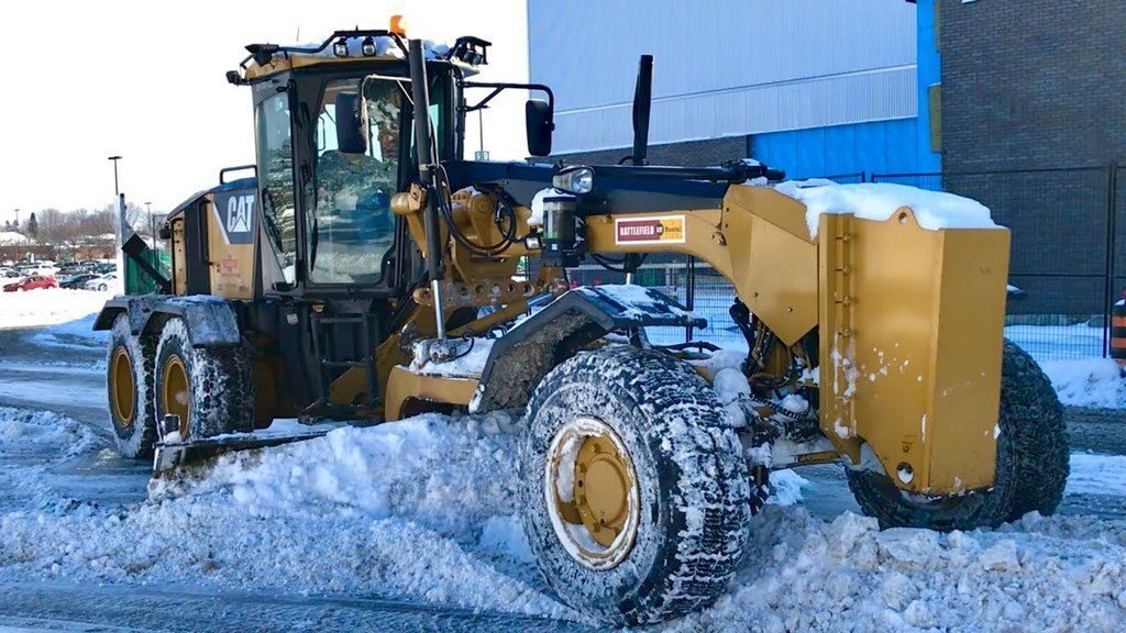 Caterpillar 160M motor grader clearing snow in a parking lot. The yellow snowplow is stopped with snow piled around its blade, and buildings are visible in the background.