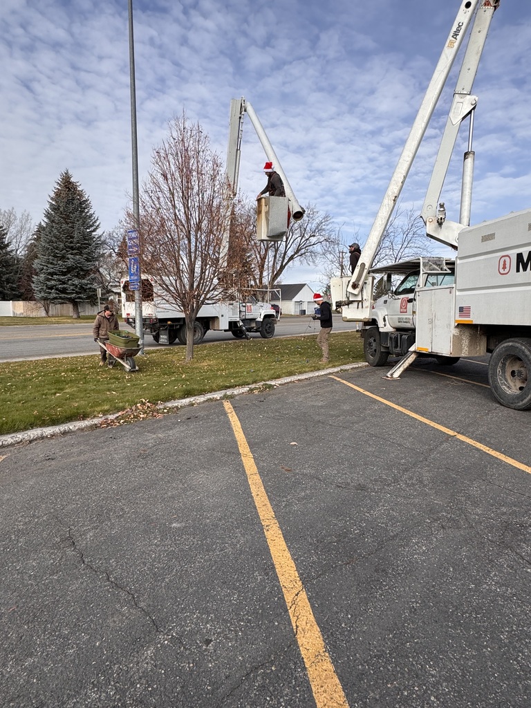 Three workers from Miller Tree Service wearing Santa hats hang Christmas lights at the City Building using two bucket trucks. One worker is elevated in a bucket near a leafless tree, another stands beside the truck, and a third pushes a wheelbarrow across the grass. The sky is partly cloudy and the scene takes place near an empty parking lot.