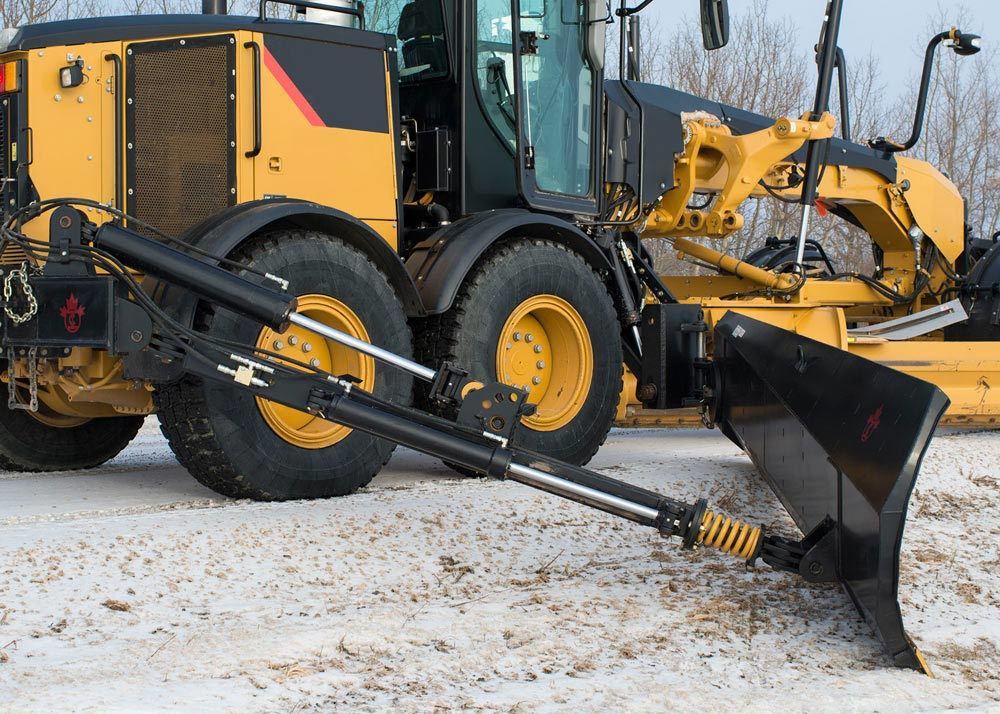 A close-up view of a yellow road grader with a hydraulic snow blade attachment angled for plowing. The machine is on a snowy roadway with bare trees in the background.