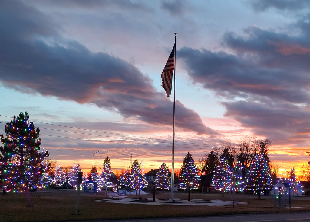 An American flag on a flagpole stands in Rotary Park at the city building at sunset, surrounded by evergreen trees wrapped in multicolored Christmas lights under a cloudy evening sky.