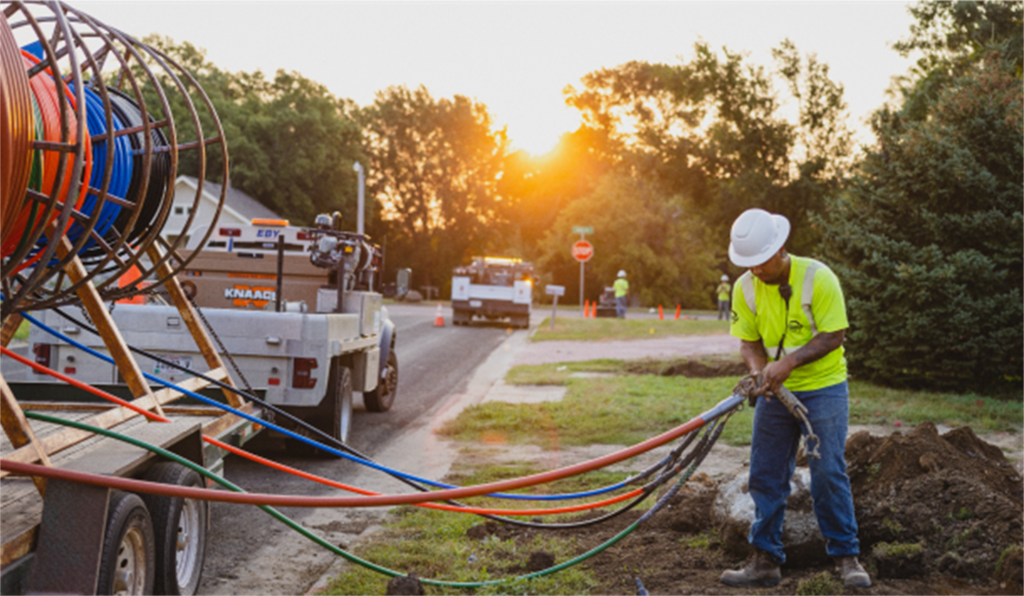 Construction crew installing underground fiber lines along a residential street, with utility trucks, marked lines, and a worker guiding colored conduit into a trench.