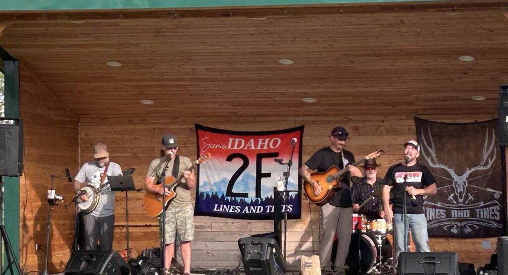 Lines and Tines performing live on an outdoor stage. Multiple musicians play guitars, banjo, drums, and vocals in front of a crowd during a community concert event.