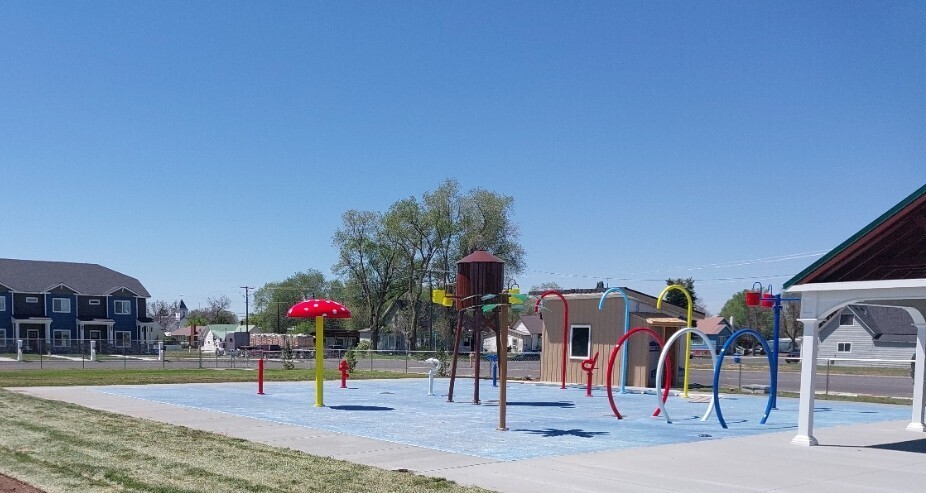 View of the St. Anthony splash pad featuring colorful water spray features, a mushroom fountain, arch sprayers, and a large tipping bucket. A pavilion is visible on the right, with homes, trees, and a blue sky in the background. The splash pad is currently closed for repairs.