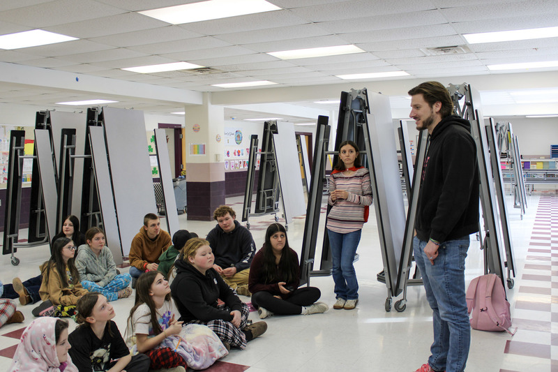 Adult male wearing a black sweatshirt speaking to a small group of younger and older students