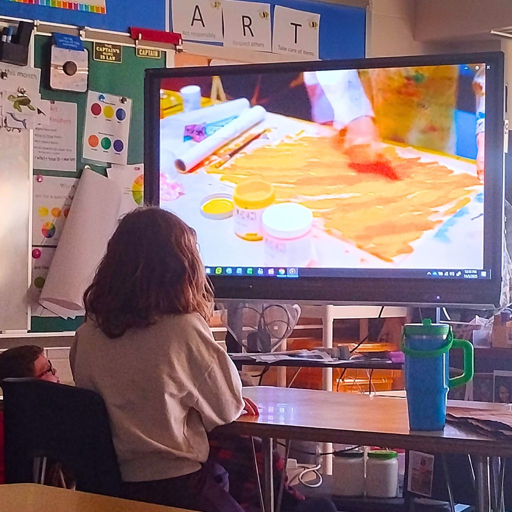 A second-grade student stands in front of a display featuring artwork by Eric Carle. The pieces are bright and colorful, showing Carle’s signature collage style with layered paper textures, bold shapes, and cheerful animal themes. The student appears curious and engaged as they observe the details.