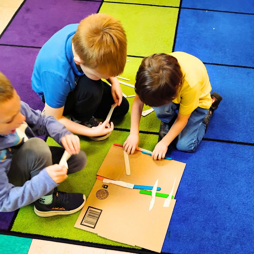 Kindergarten students are gathered around tables working together on a building project. They’re using materials like paper, cardboard, straws, and tape to design small houses inspired by The Three Little Pigs. The classroom looks bright and colorful, with students smiling and talking as they build.
