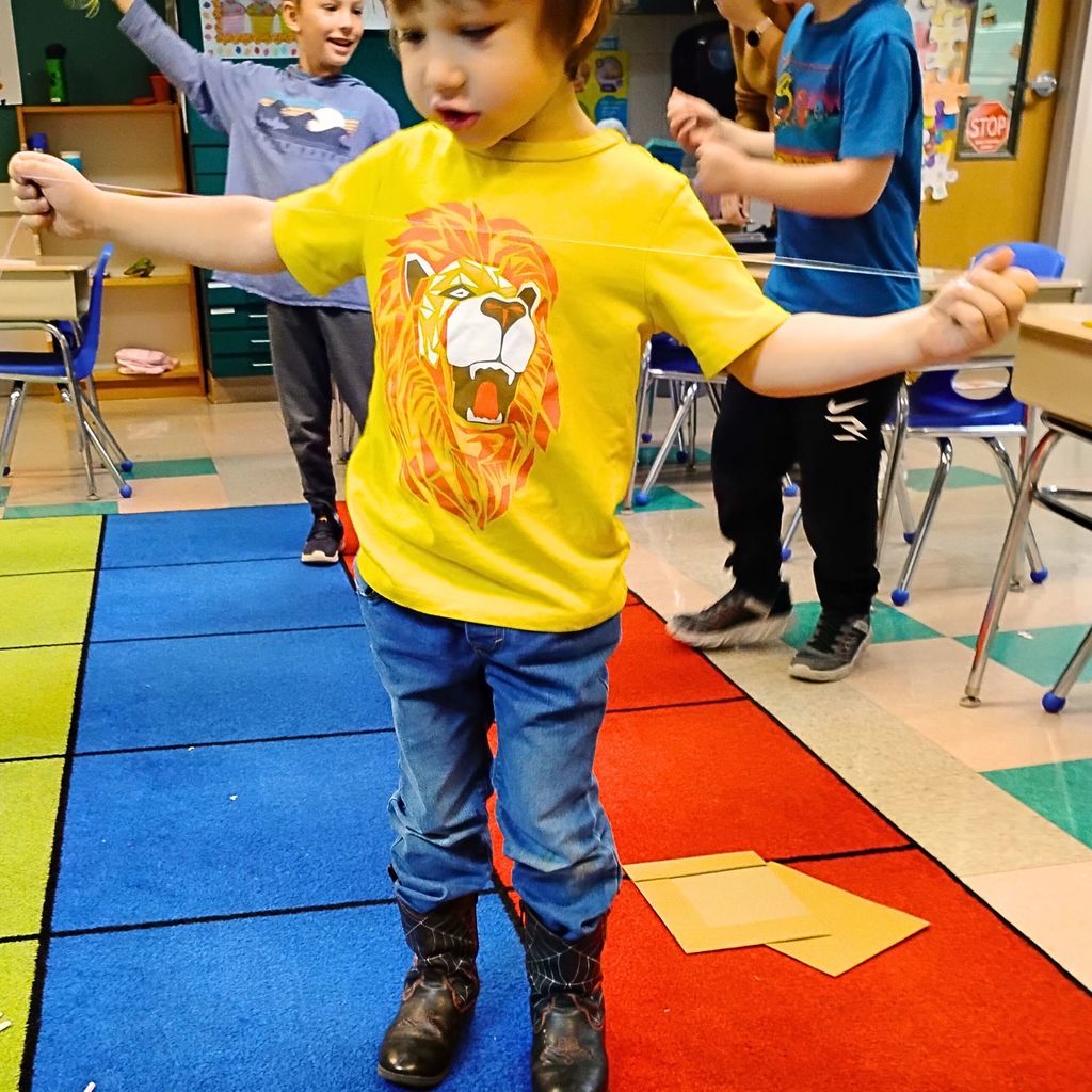 Kindergarten students are gathered around tables working together on a building project. They’re using materials like paper, cardboard, straws, and tape to design small houses inspired by The Three Little Pigs. The classroom looks bright and colorful, with students smiling and talking as they build.