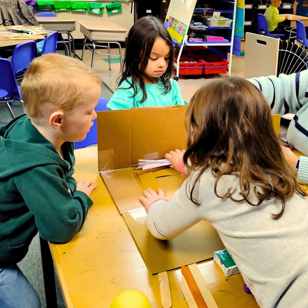 Kindergarten students are gathered around tables working together on a building project. They’re using materials like paper, cardboard, straws, and tape to design small houses inspired by The Three Little Pigs. The classroom looks bright and colorful, with students smiling and talking as they build.
