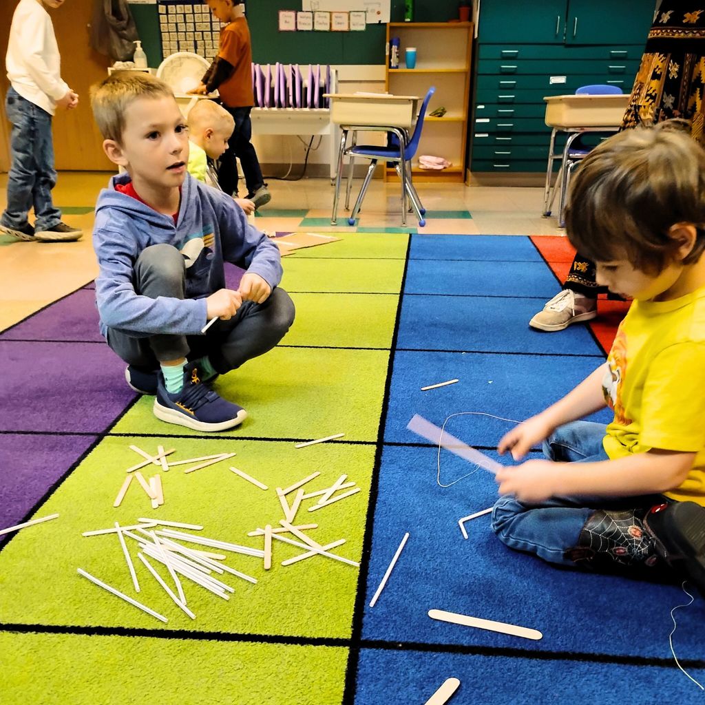 Kindergarten students are gathered around tables working together on a building project. They’re using materials like paper, cardboard, straws, and tape to design small houses inspired by The Three Little Pigs. The classroom looks bright and colorful, with students smiling and talking as they build.