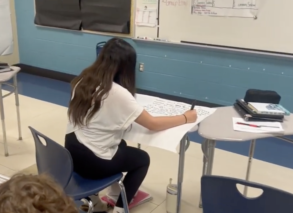 Student is sitting a desk in the clasrrom writing on an oversize piece of paper.