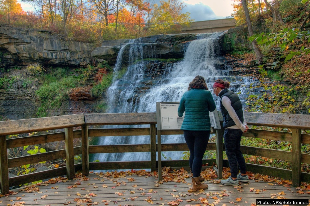 Two visitors stand on a wooden observation deck reading an interpretive sign at Brandywine Falls in Cuyahoga Valley National Park. The multi-tiered waterfall cascades over layered shale rock in the background, surrounded by trees in peak autumn color. Fallen leaves cover the deck floor. Photo by Bob Trinnes/NPS.