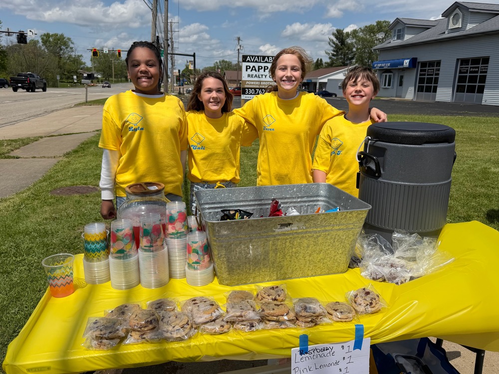Four fourth-grade students in matching yellow lemonade-themed t-shirts smile behind their stand, which is stocked with chocolate chip cookies, cups, and a large drink cooler. A handwritten sign advertises raspberry lemonade, pink lemonade, and cookies for $1 each.