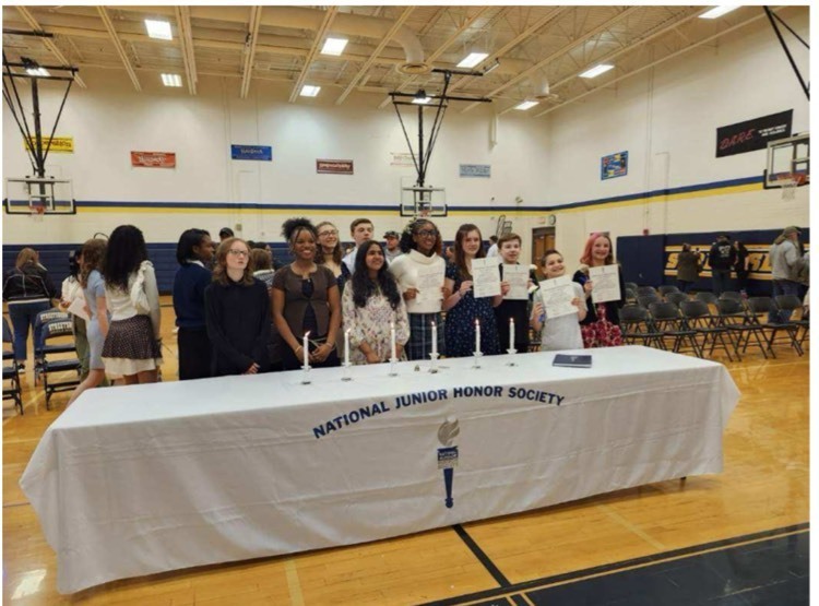 students stand in front of a table with a white tablecloth that says national junior honor society in what looks like a gym  