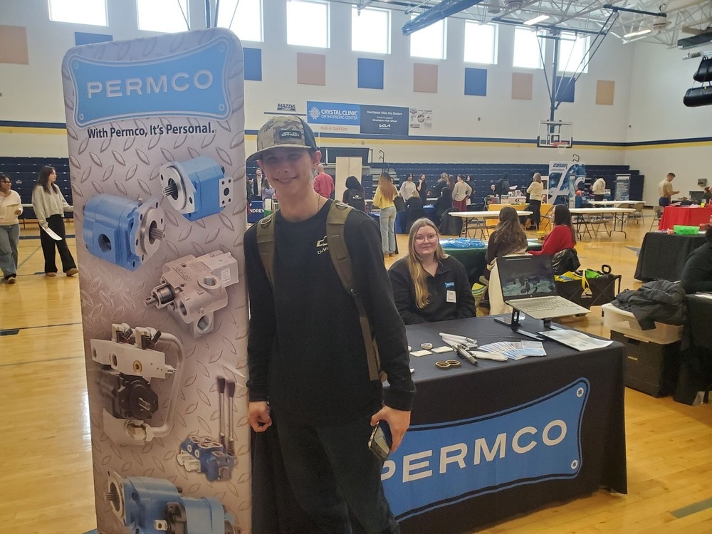 A student stops at the Permco booth during a career fair held in a school gymnasium. A company representative sits behind a branded table display as other students and employers interact in the background.
