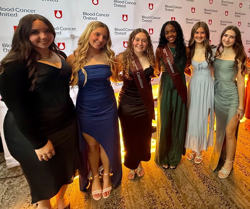 Six young women in formal gowns pose together in front of a Blood Cancer United step-and-repeat backdrop. Two of the women wear maroon "Candidate" sashes, indicating their participation in a Blood Cancer United fundraising campaign.