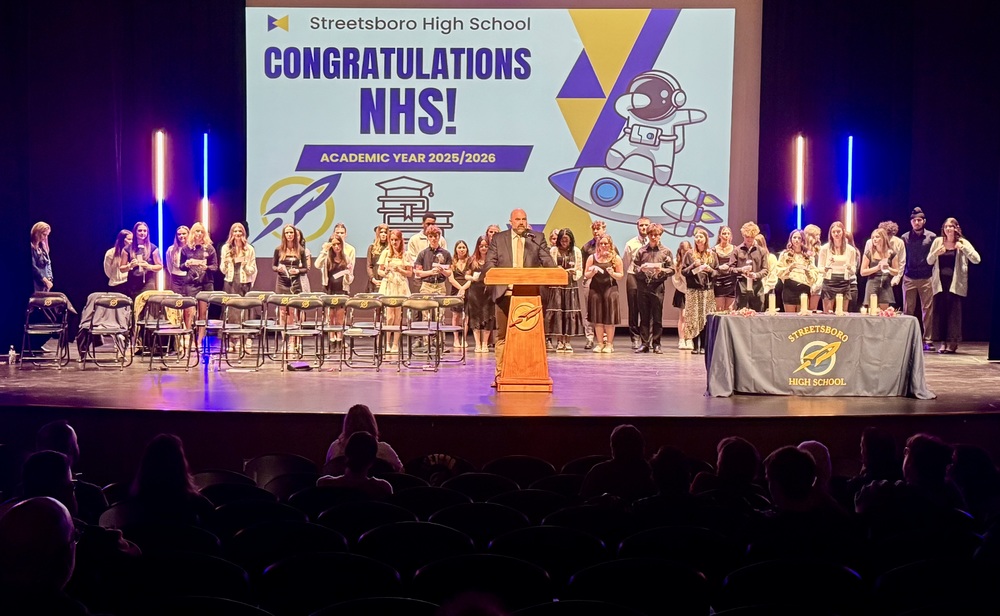 Students stand on stage behind chairs. Behind them is a sign that says "Congratualtions NHS"