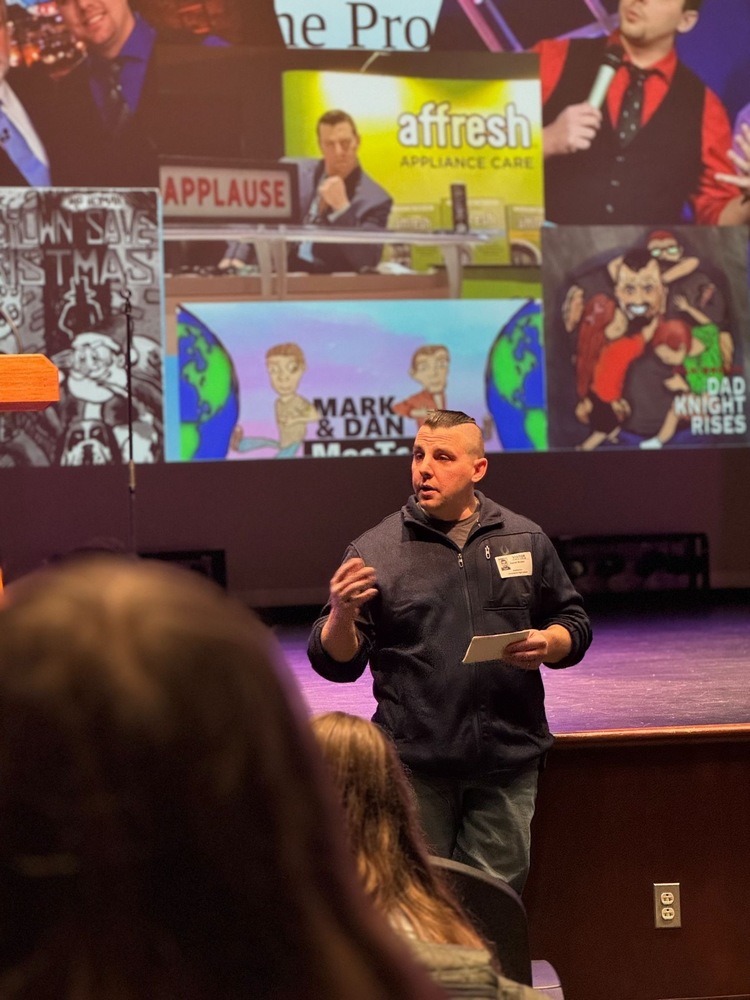 Dan Brown standing on the stage in the SPAC with photos of himself on the screen behind him.