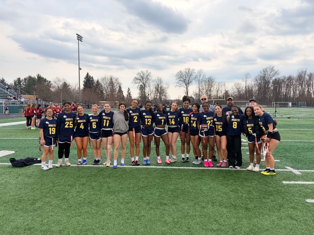 Alt text: A girls' flag football team called the Rockets poses together on a green turf field after a game. The players wear matching navy blue and gold uniforms with jersey numbers ranging from 1 to 25. Three coaches stand at the right end of the group. The background shows bleachers, bare trees, a light tower, and an overcast sky.