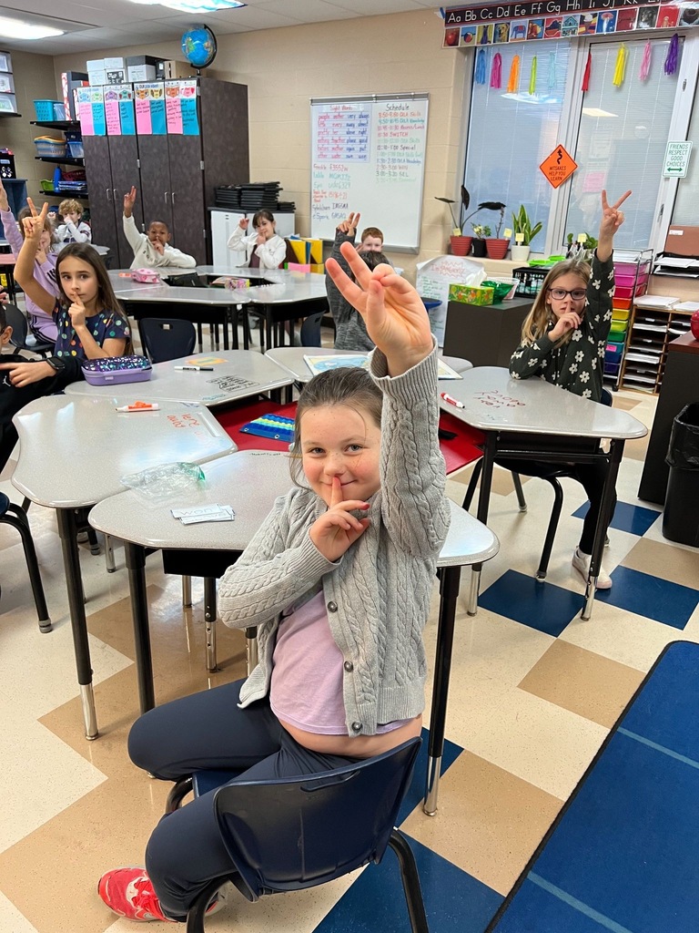 SES 2Close-up of a smiling SES student in a gray cardigan demonstrating the PAX Quiet signal with two fingers raised while sitting at her desk, with classmates visible in the background also practicing the routine.