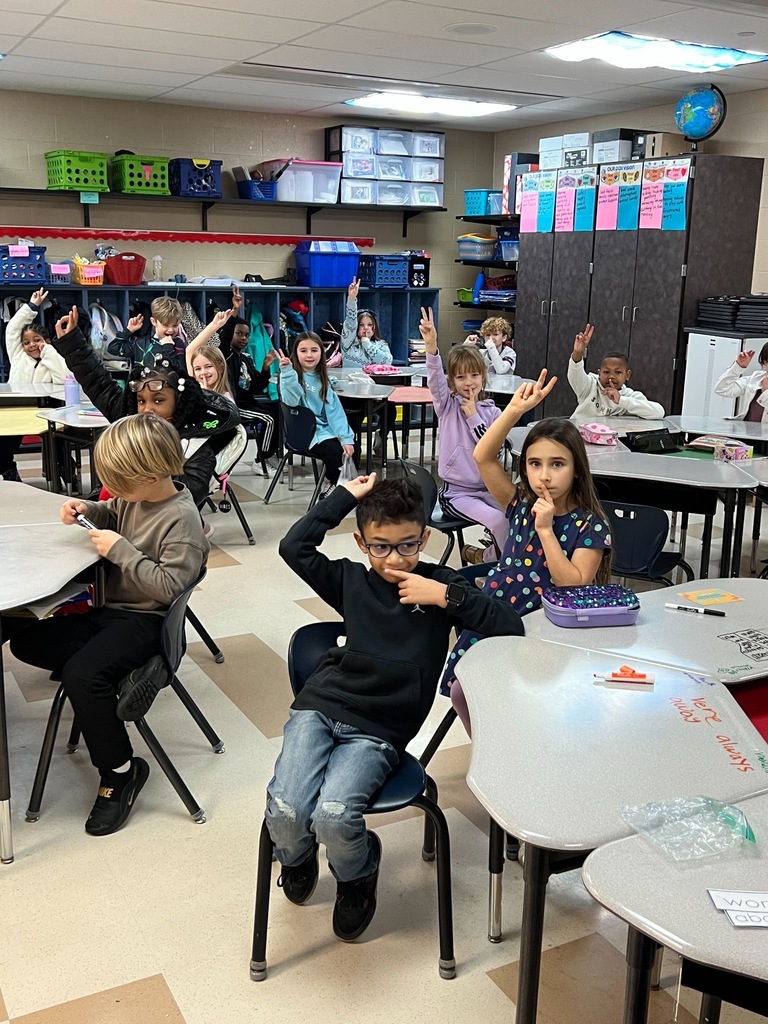 SES students in a classroom practicing PAX Quiet hand signal with two fingers raised, sitting at tables with learning materials visible on shelves in the background.