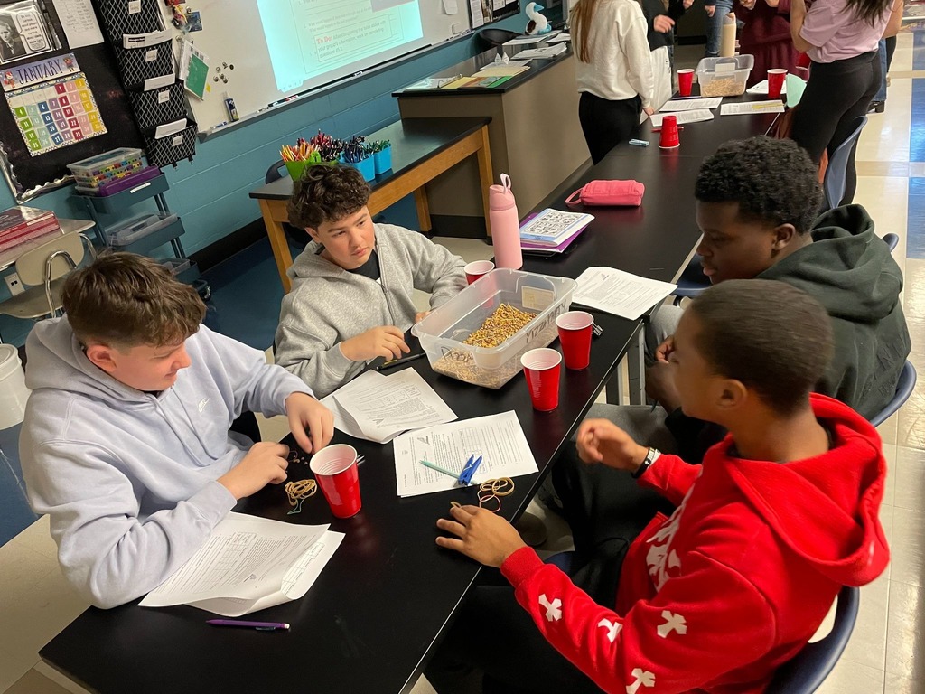 Students standing around tables in a classroom working on hands-on projects in groups. 