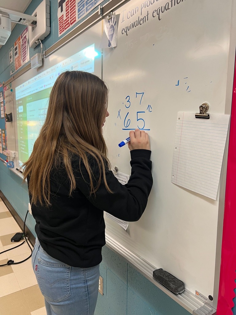 tudent in black sweatshirt and jeans working on a long division math problem at the whiteboard, solving 37 divided by 65. Classroom setting with projector screen and educational posters visible in background.