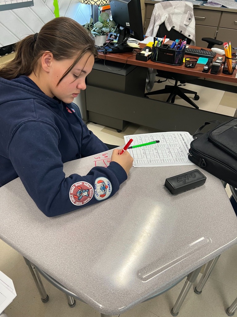 student wearing navy blue school sweatshirt with rocket logo working on a math worksheet at desk, using red and green markers. Classroom setting with teacher's desk visible in background.