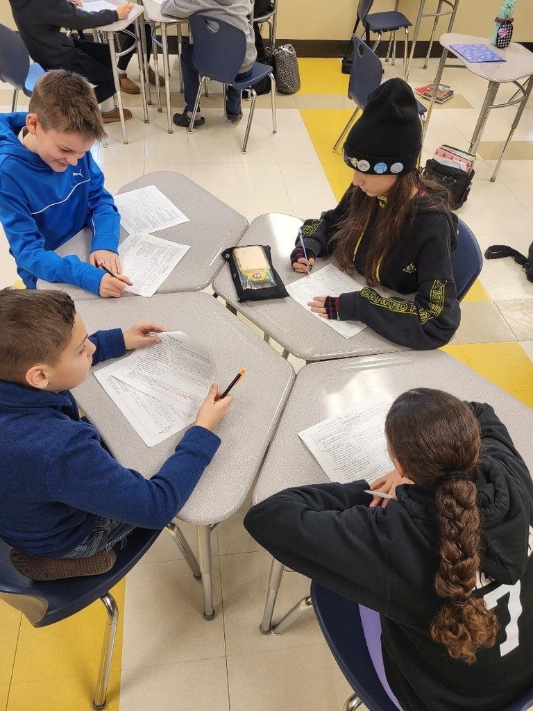 four students collaborating at desks arranged in a pod, working on reading assignments with pencils and papers. Students wearing casual clothing including blue and black sweatshirts. Bright yellow floor stripe visible between classroom desks.