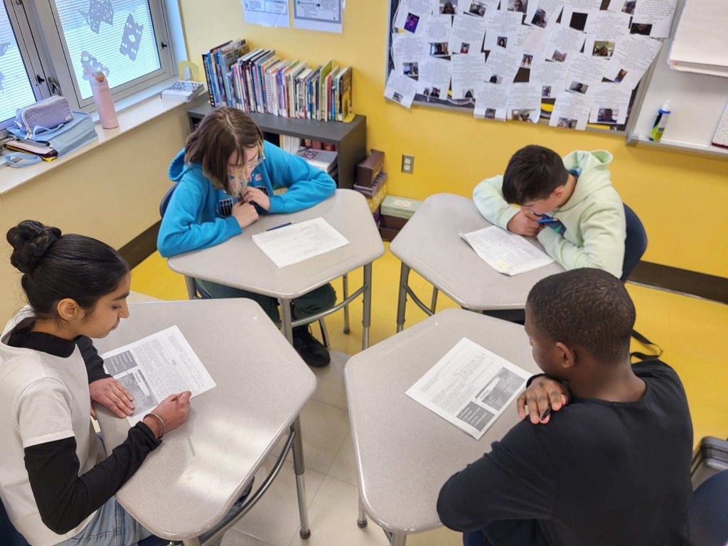four students working independently on reading assignments at individual desks arranged in a pod formation. Bright yellow classroom wall with bulletin board displaying student photos and work. Bookshelf and windows visible in background.