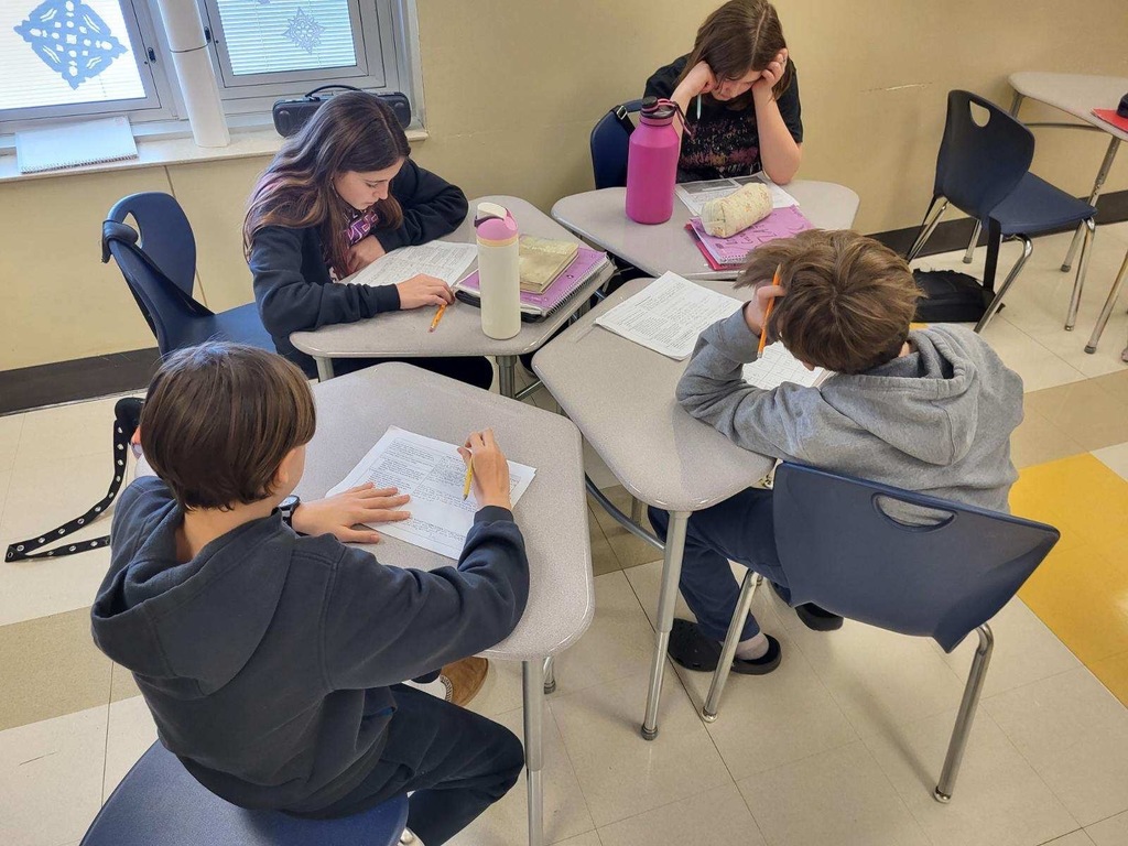 four students working independently on written assignments at individual desks with papers, notebooks, and water bottles. Students wearing casual sweatshirts and hoodies in classroom with windows and snowflake decorations visible in background.