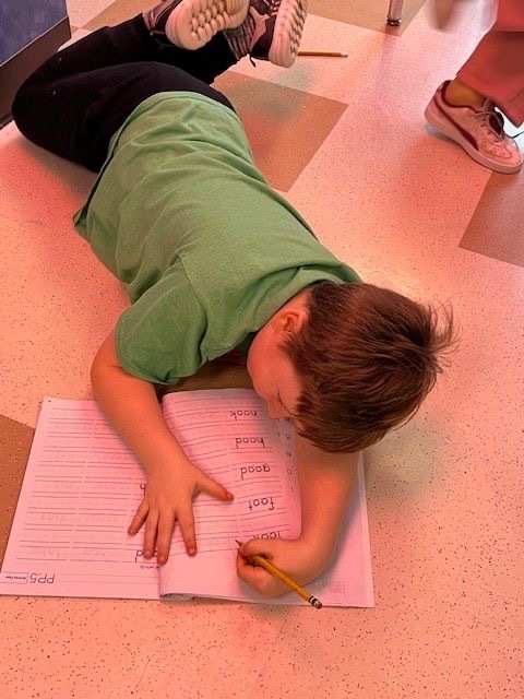 elementary student in green shirt lying on classroom floor writing spelling words on pink lined paper with pencil. Other students' feet visible in background on speckled tile floor.