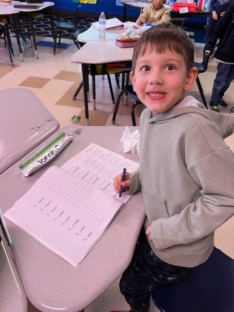 smiling elementary student in gray hoodie practicing rainbow writing at desk with pencil and worksheet, using EXPO dry erase markers. Classroom setting with other students working at tables in background.