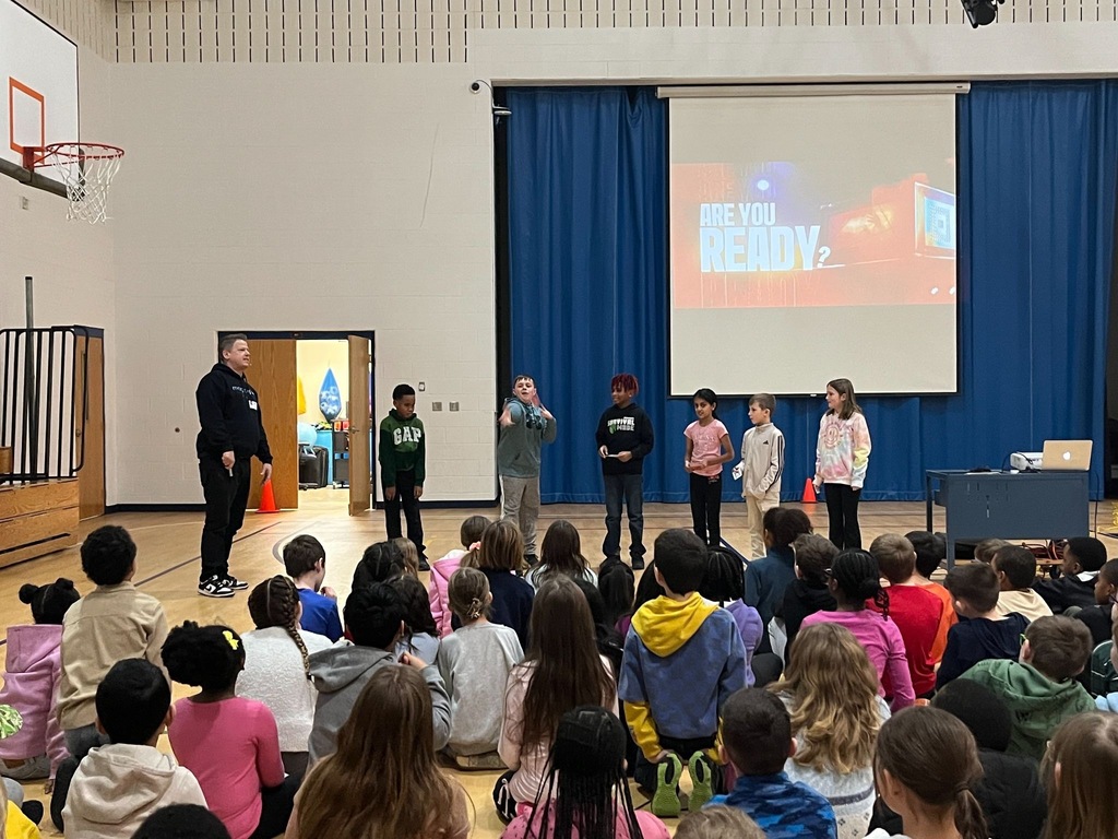Students sitting on the gym floor for an assembly There is a row of other students standing in front of them with a projection screen behind them that says in white letter: "Are you ready?"