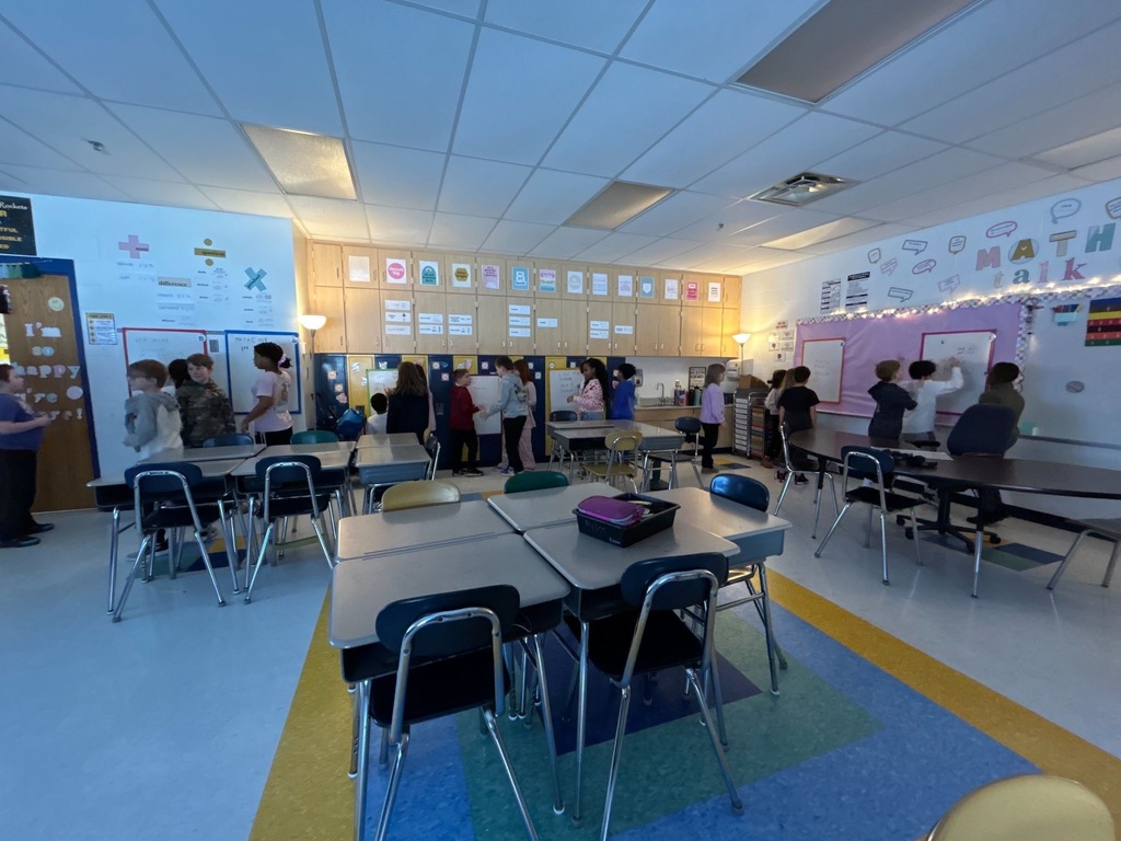 Students standing around white boards in the background of a classroom. Empty desks are in the forefront of the photo as all the students are standing around in groups around whiteboards.