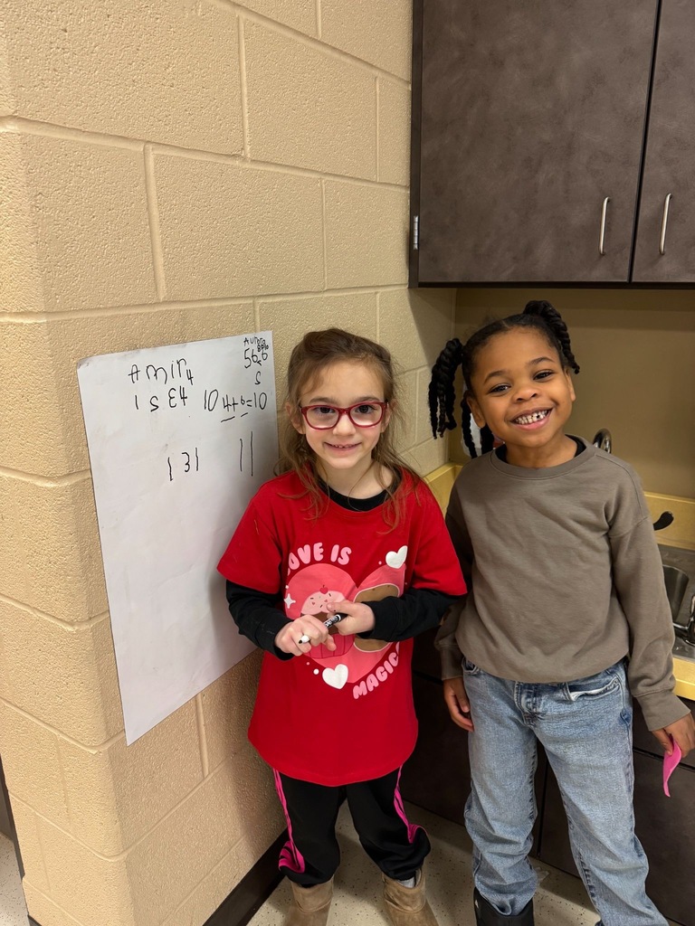 Two students standing on the right side of a vertical whiteboard. With their names and math problems written on the board.