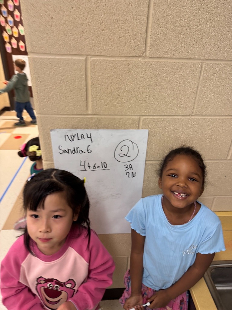 Two students standing on either side of a vertical whiteboard. With their names and math problems written on the board.