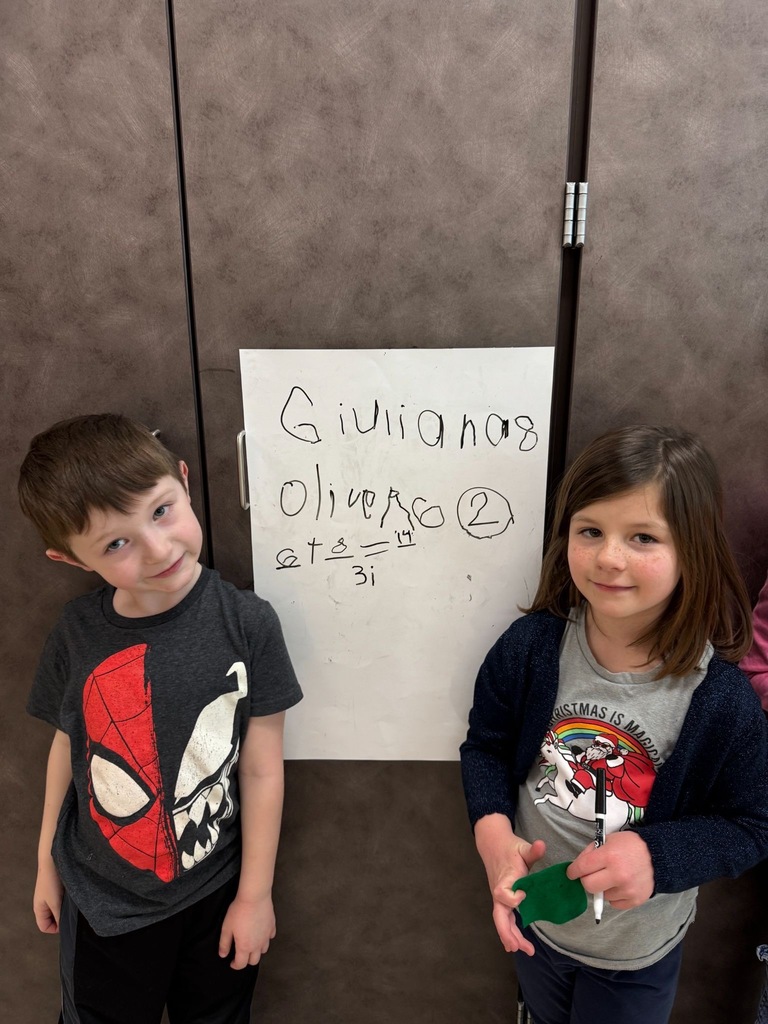 Two students standing on either side of a vertical whiteboard. With their names and math problems written on the board.