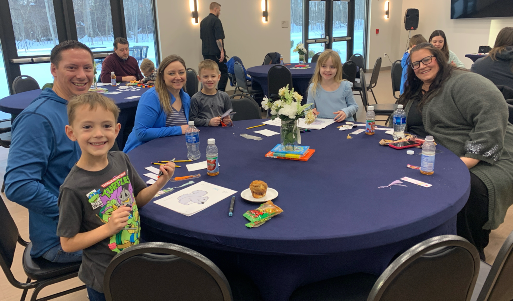 Families sitting around tables with breakfast items and coloring pages on the tables in front of them