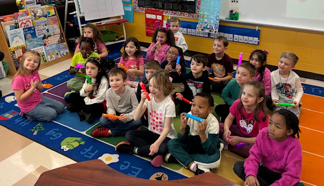 "A kindergarten class of approximately 20 students sits together on a colorful alphabet rug in their classroom, each holding brightly colored toy harmonicas or musical instruments. Many students are wearing pink and red clothing. The classroom features a bookshelf, bulletin boards, and educational displays on the yellow walls behind them. The children are smiling and looking toward the camera.