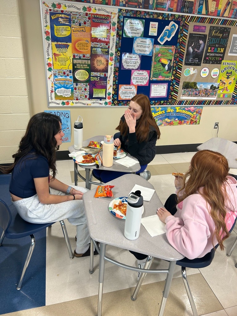 Students sitting around desks eating pizza in a classroom setting. 