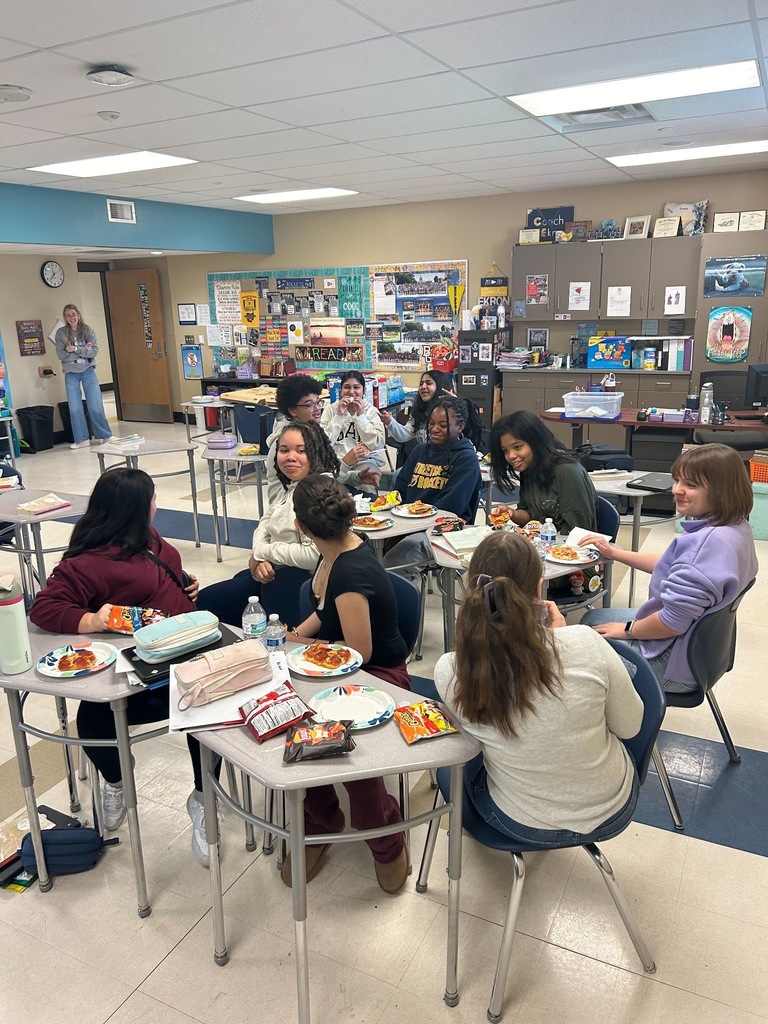 Students sitting around desks eating pizza in a classroom setting. 