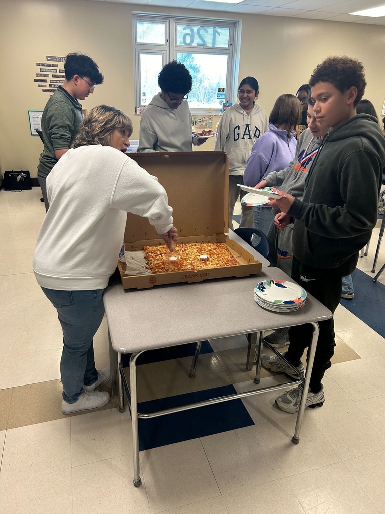Students sitting around desks eating pizza in a classroom setting. 