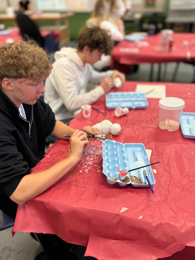 Student holding brushes and sitting at a covered table working on art project.