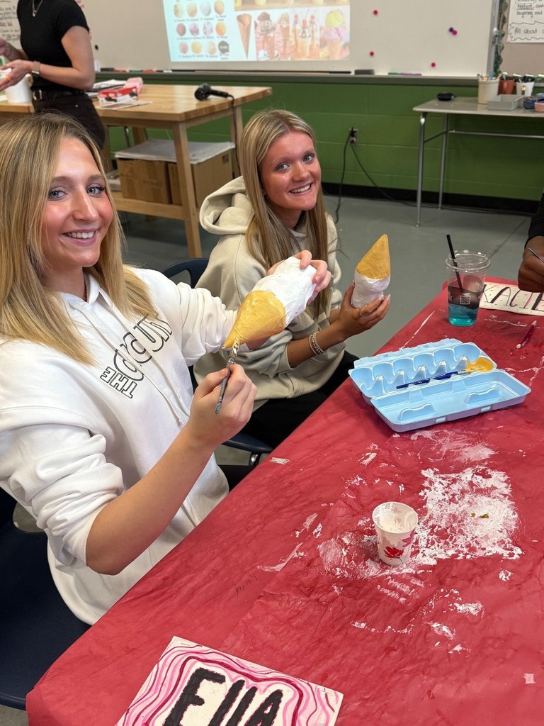 Students holding brushes and sitting at a covered table working on art project.
