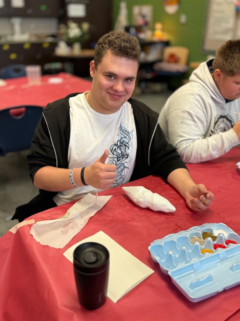 Student holding brushes and sitting at a covered table working on art project.