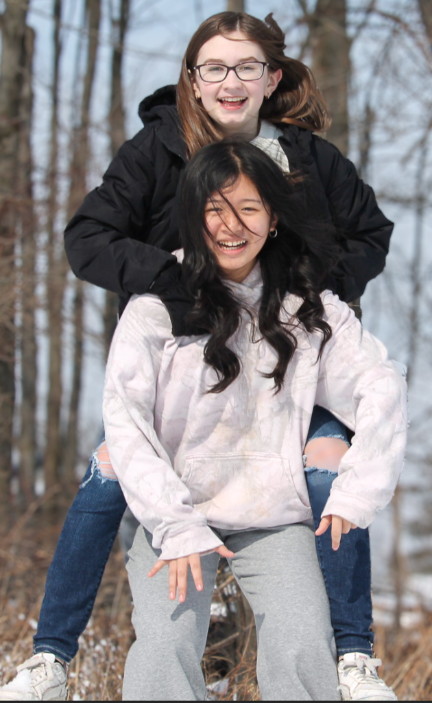 Two teenage girls laugh outdoors on a winter day, one giving the other a piggyback ride, with bare trees and snow visible in the background.