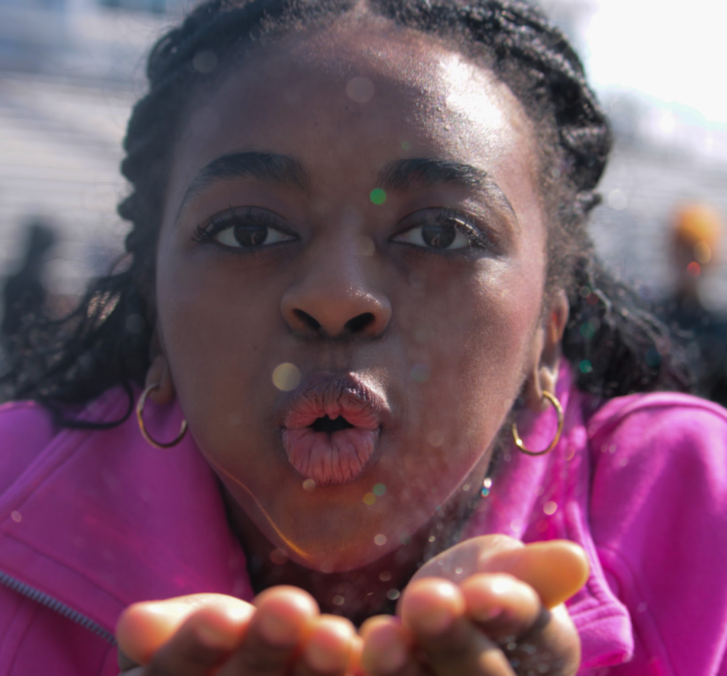 A woman in a bright pink jacket blows glitter from her cupped hands toward the camera, with colorful sparkles visible in the air around her face.
