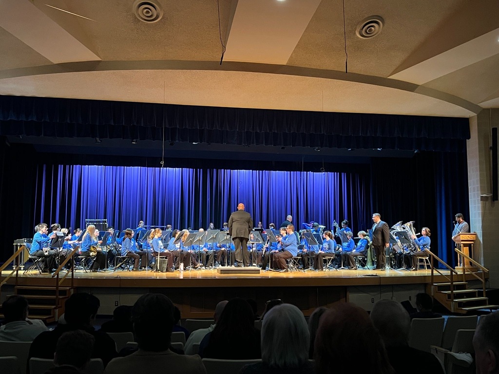 A student band in blue shirts performs on a school auditorium stage under blue lighting, with a conductor leading them before an audience.unknown 2