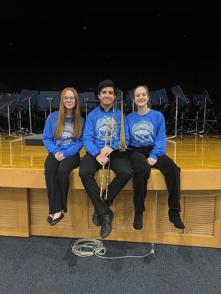Three students in matching blue American School Band Directors Association shirts sit on the edge of a stage, the middle student holding a trombone.